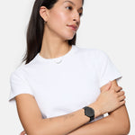 Eine Frau in einem weißen T-Shirt mit dem Tamaris Set - The Sparkling Black von Tamaris, bestehend aus einer schwarzen Uhr und einem Glitzerarmband, verschränkt die Arme und blickt zur Seite - eine elegante Accessoire-Kombination für den täglichen Stil.
