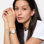 Eine Frau mit langen dunklen Haaren und weißem Hemd zeigt ihre Tamaris Armbanduhr - The Elegance mit funkelnden Zirkoniasteinen und einem eleganten Edelstahlband, während sie ihr Kinn sanft auf ihre Hände stützt.