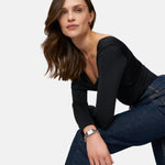 Eine Frau in einem schwarzen langärmeligen Oberteil und Jeans posiert selbstbewusst und hebt die Tamaris Armbanduhr - Chain Watch von Tamaris hervor, während sie ihren Arm auf ihr Knie vor einem weißen Hintergrund stützt.