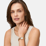 Frau mit hellbraunem Haar, die ein beiges Tank-Top und eine Tamaris Armbanduhr - Chain Watch von Tamaris trägt, berührt ihren Hals und schaut nachdenklich in die Kamera.