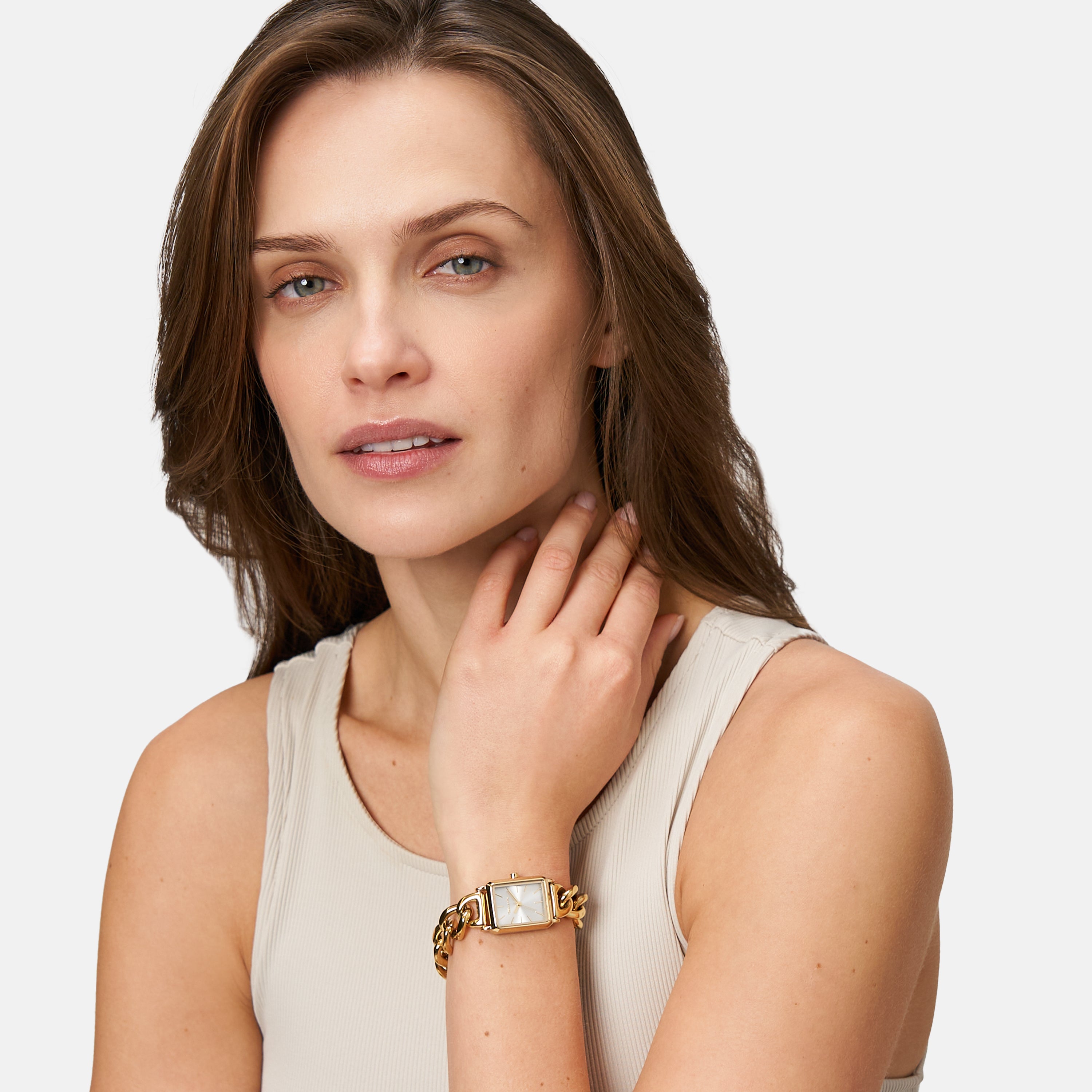 Frau mit hellbraunem Haar, die ein beiges Tank-Top und eine Tamaris Armbanduhr - Chain Watch von Tamaris trägt, berührt ihren Hals und schaut nachdenklich in die Kamera.