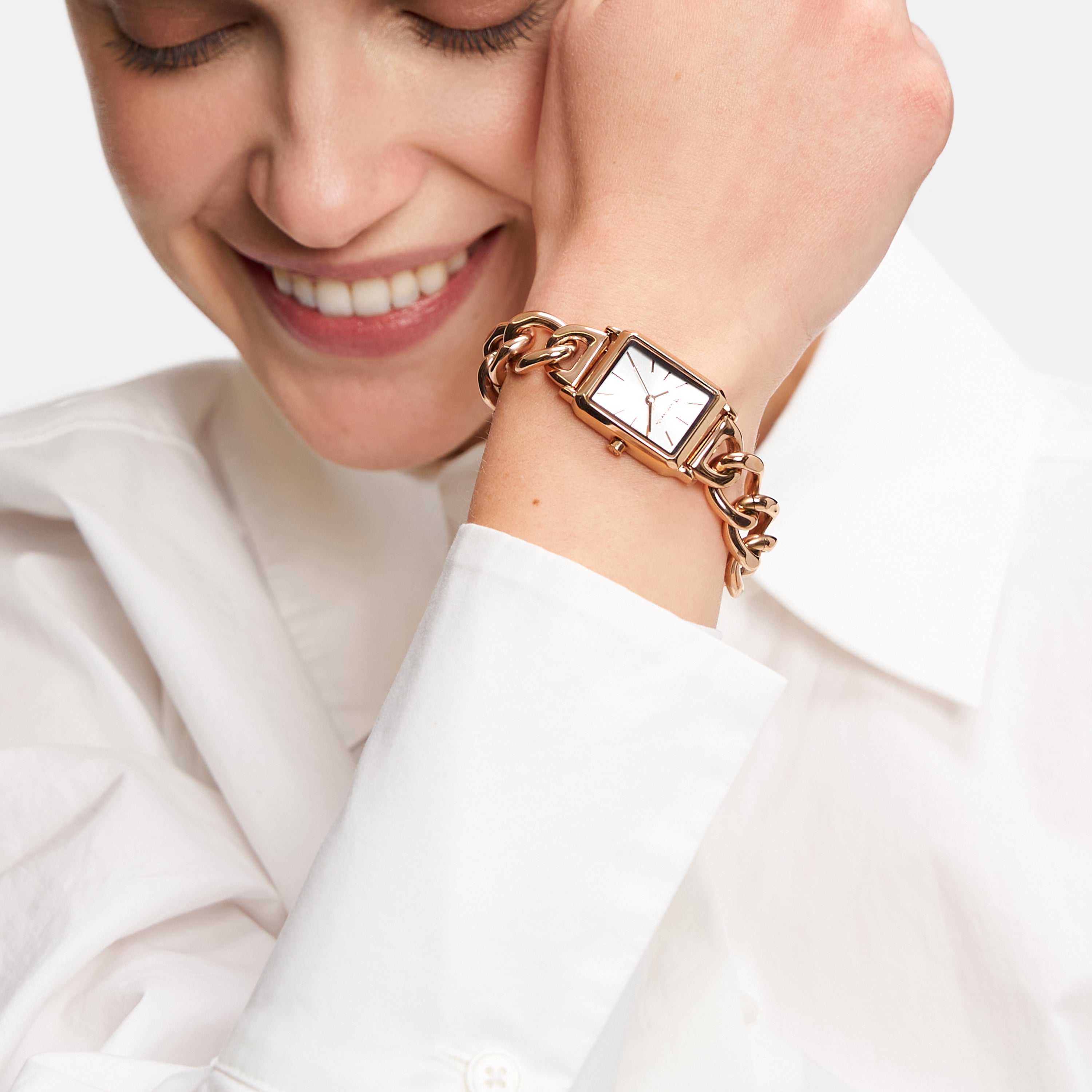 Lächelnde Frau in weißem Hemd trägt eine Tamaris Armbanduhr - Chain Watch von Tamaris mit einem goldenen Kettenband und einem eleganten rechteckigen Mini-Zifferblatt an ihrem Handgelenk.