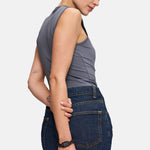 Eine Frau in einem grauen, ärmellosen Oberteil und einem dunklen Jeansrock steht seitlich und blickt mit dem Arm hinter dem Rücken nach unten, wobei sie eine Tamaris Armbanduhr - Chain Watch von Tamaris zierlich zur Schau stellt.