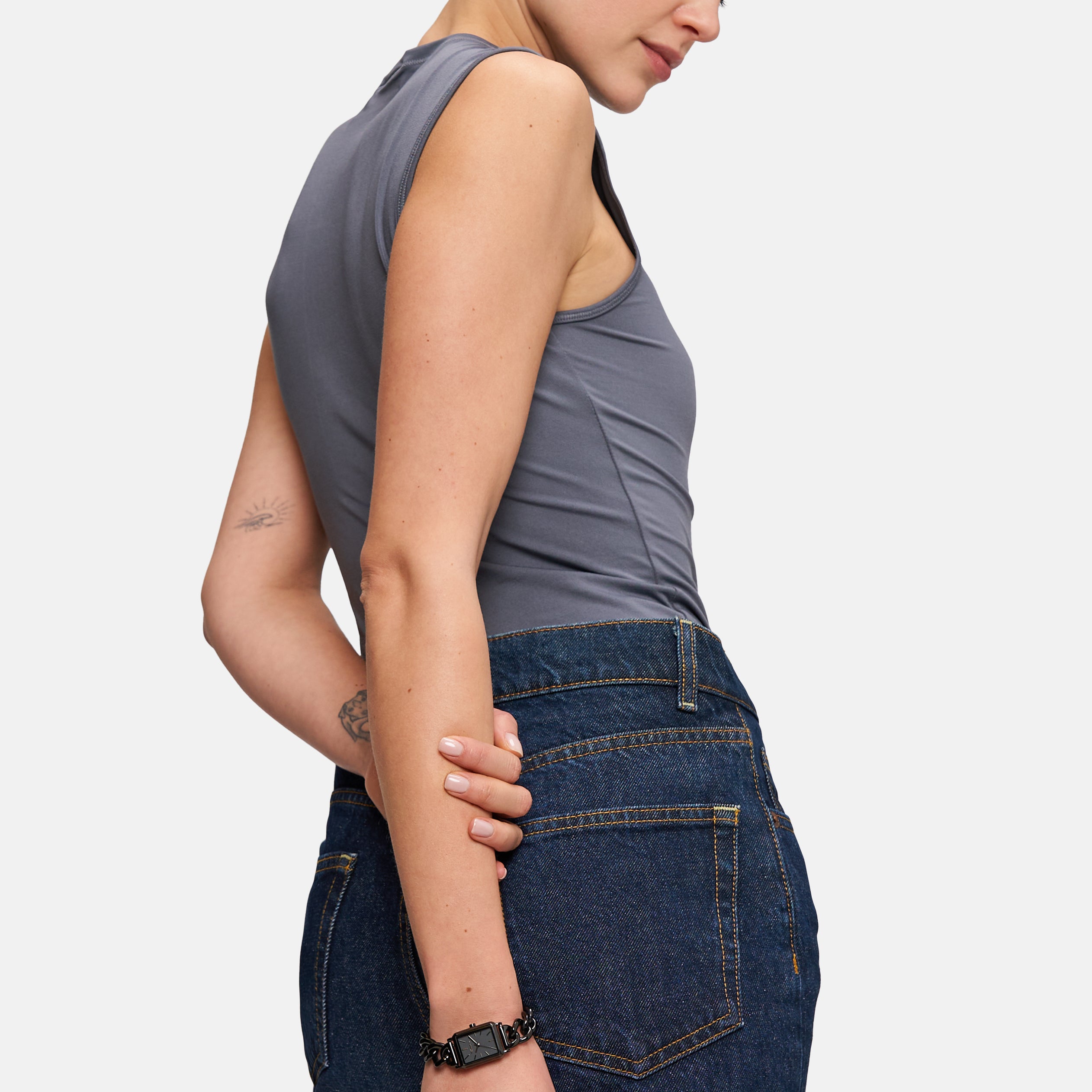 Eine Frau in einem grauen, ärmellosen Oberteil und einem dunklen Jeansrock steht seitlich und blickt mit dem Arm hinter dem Rücken nach unten, wobei sie eine Tamaris Armbanduhr - Chain Watch von Tamaris zierlich zur Schau stellt.