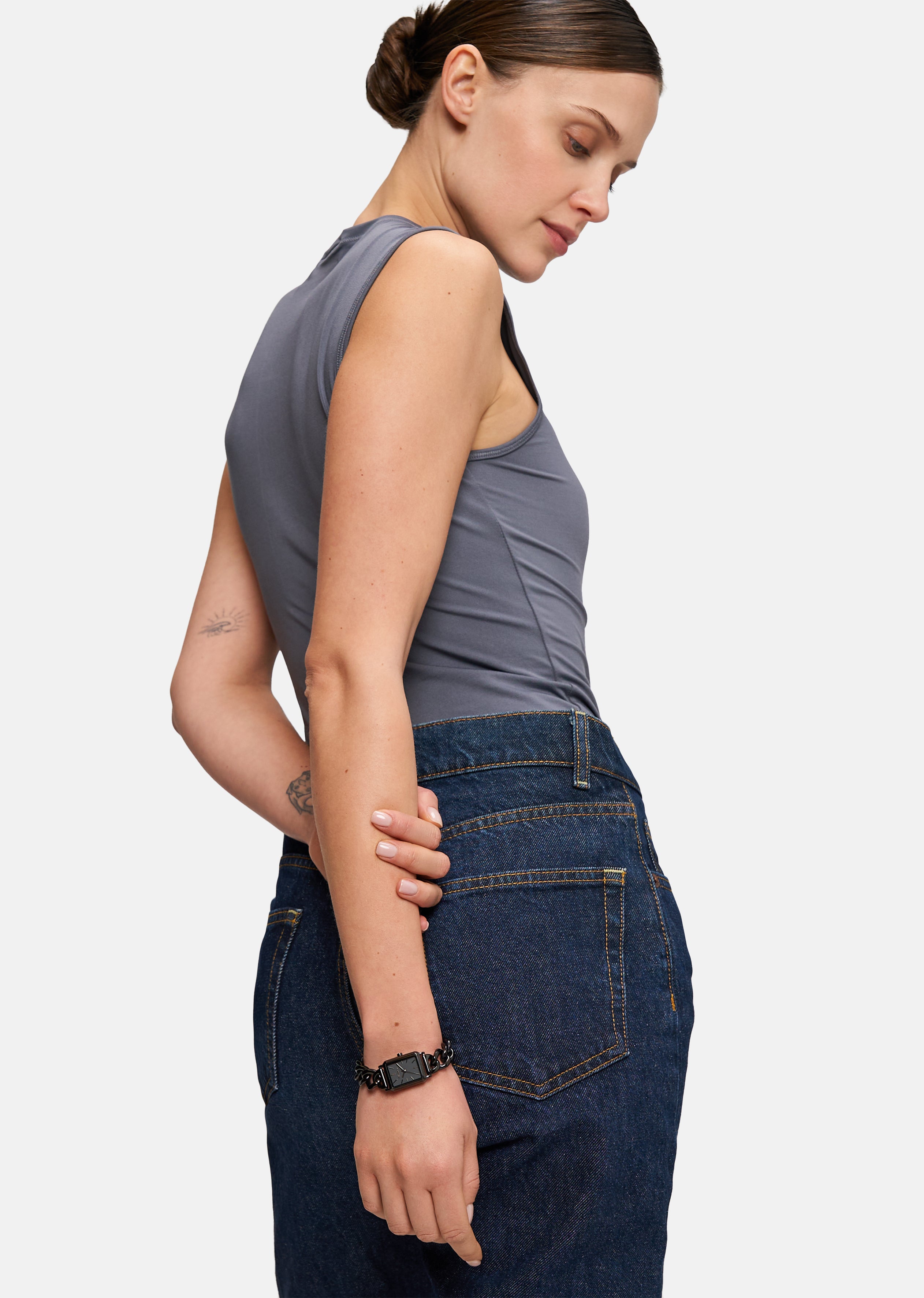 Eine Frau in einem grauen, ärmellosen Oberteil und einem dunklen Jeansrock steht seitlich und blickt mit dem Arm hinter dem Rücken nach unten, wobei sie eine Tamaris Armbanduhr - Chain Watch von Tamaris zierlich zur Schau stellt.