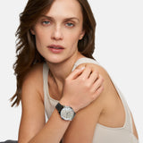 Eine Frau in einem beigen Tank-Top modelliert die Tamaris Armbanduhr - Basic Elegance mit einem eleganten Perlmuttzifferblatt und blickt in die Kamera vor einem schlichten Hintergrund.