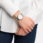 Eine Person in weißem Hemd und Jeans trägt die stilvolle Tamaris Armbanduhr - Clover, eine silberne Tamaris-Uhr mit weißem Zifferblatt und elegantem Netzband.