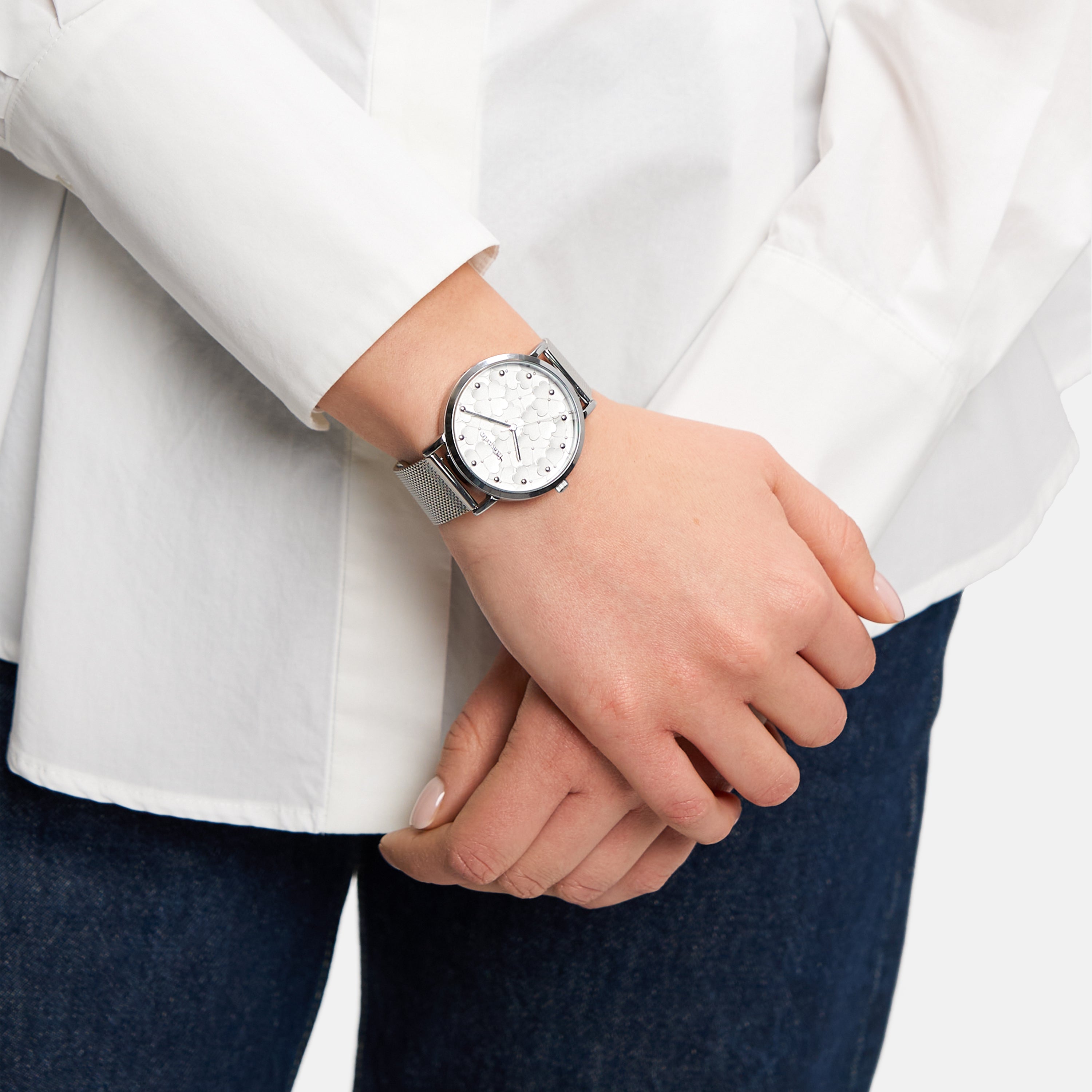 Eine Person in weißem Hemd und Jeans trägt die stilvolle Tamaris Armbanduhr - Clover, eine silberne Tamaris-Uhr mit weißem Zifferblatt und elegantem Netzband.