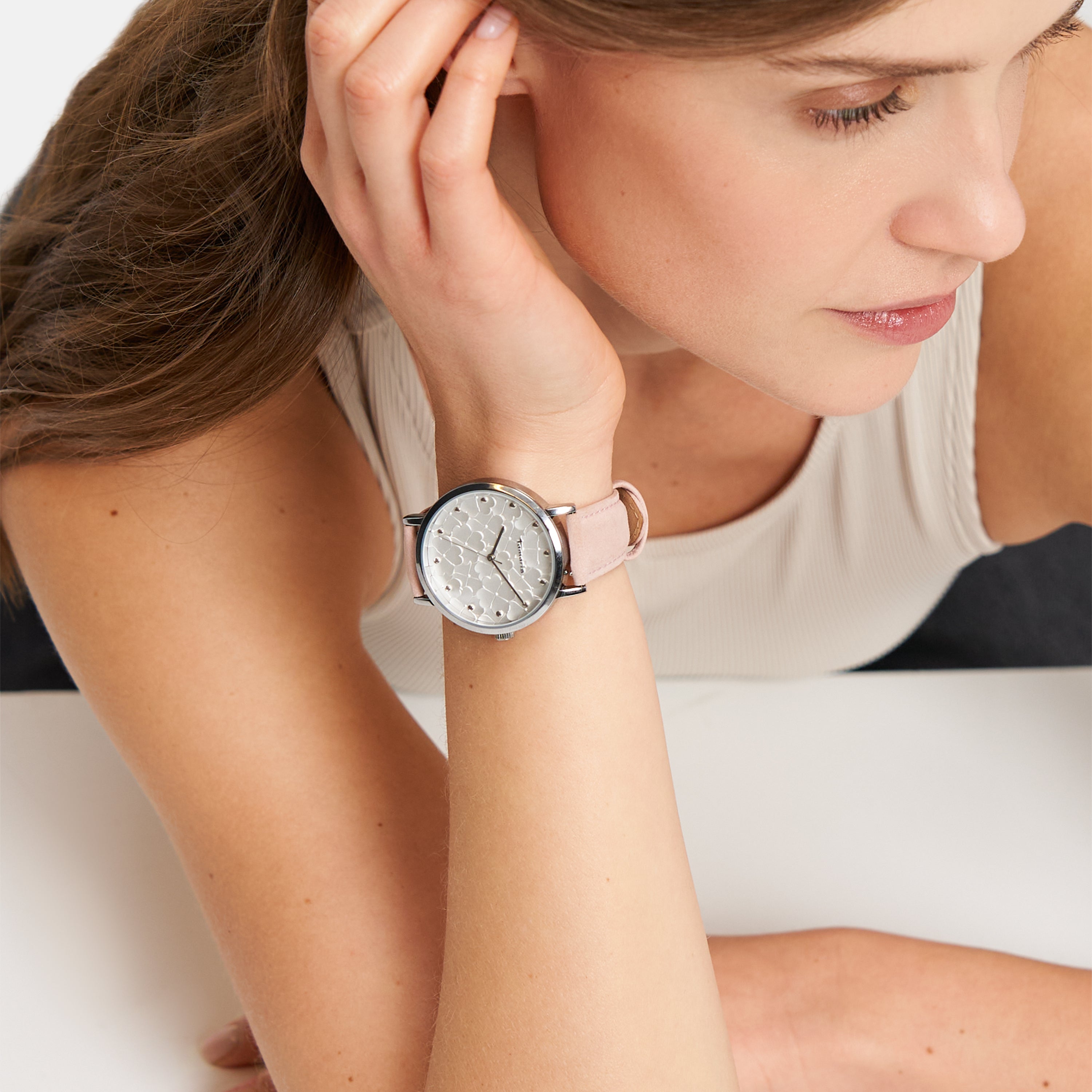 Eine Frau in einem ärmellosen Top stützt ihren Kopf auf die Hand und trägt die Tamaris Armbanduhr - Clover mit floralem Zifferblatt und stilvollem rosa Lederband von Tamaris am Handgelenk.