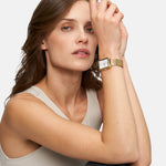 Eine Frau in einem ärmellosen Oberteil trägt die Tamaris Armbanduhr - Square Meets Mesh mit einem goldenen Netzband, während sie ihren Arm auf ihr Knie stützt und in die Kamera schaut.