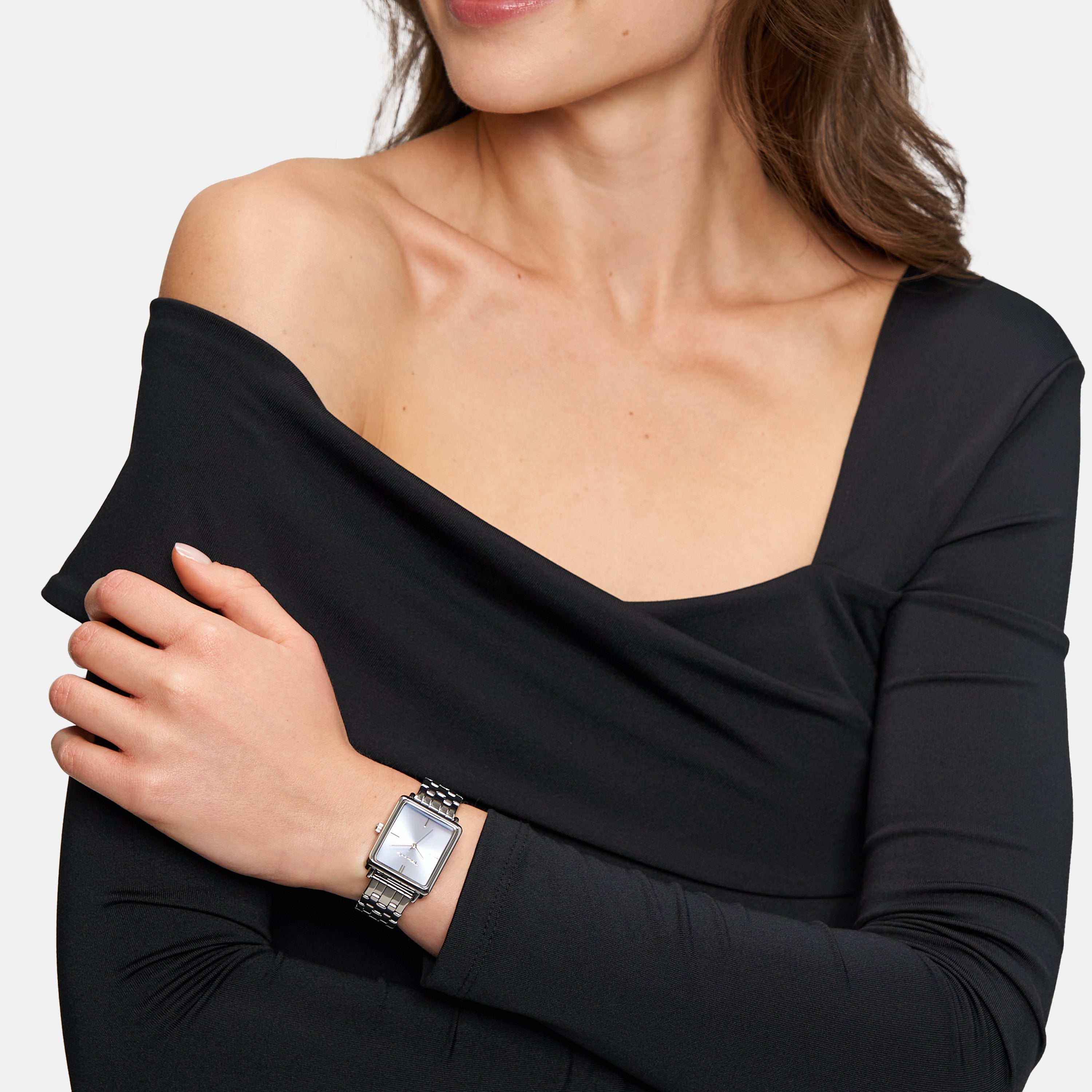 Eine Frau in einem schwarzen schulterfreien Top, die Arme verschränkt und leicht lächelnd, trägt die Tamaris Armbanduhr - The Bicolor Square mit einem schlanken quadratischen Gehäuse und einem eleganten Edelstahlband.