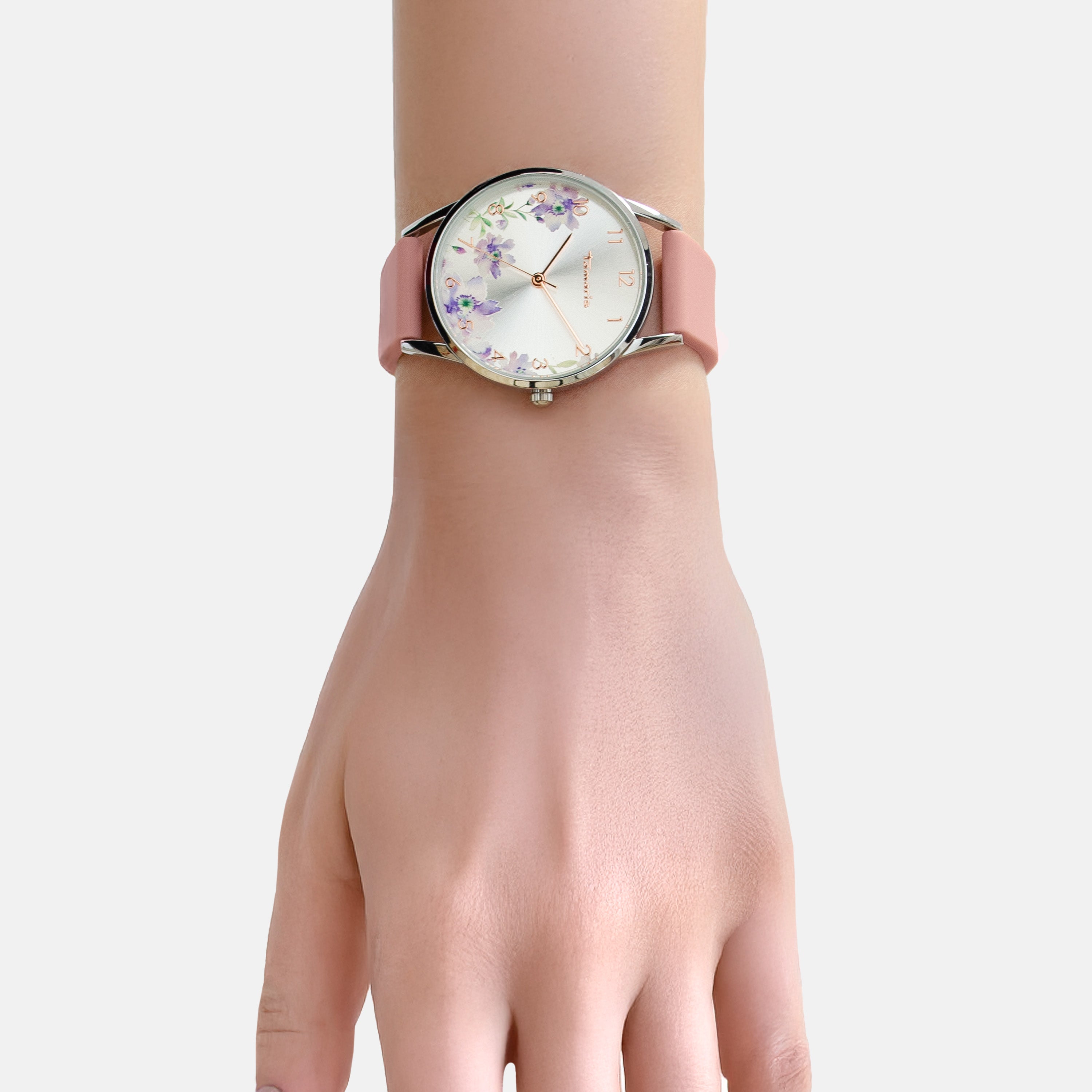 Eine Hand trägt die Tamaris Armbanduhr - The Flower von Tamaris mit einem rosafarbenen Silikonarmband und einem floralen Ziffernblattdesign vor einem hellen Hintergrund.
