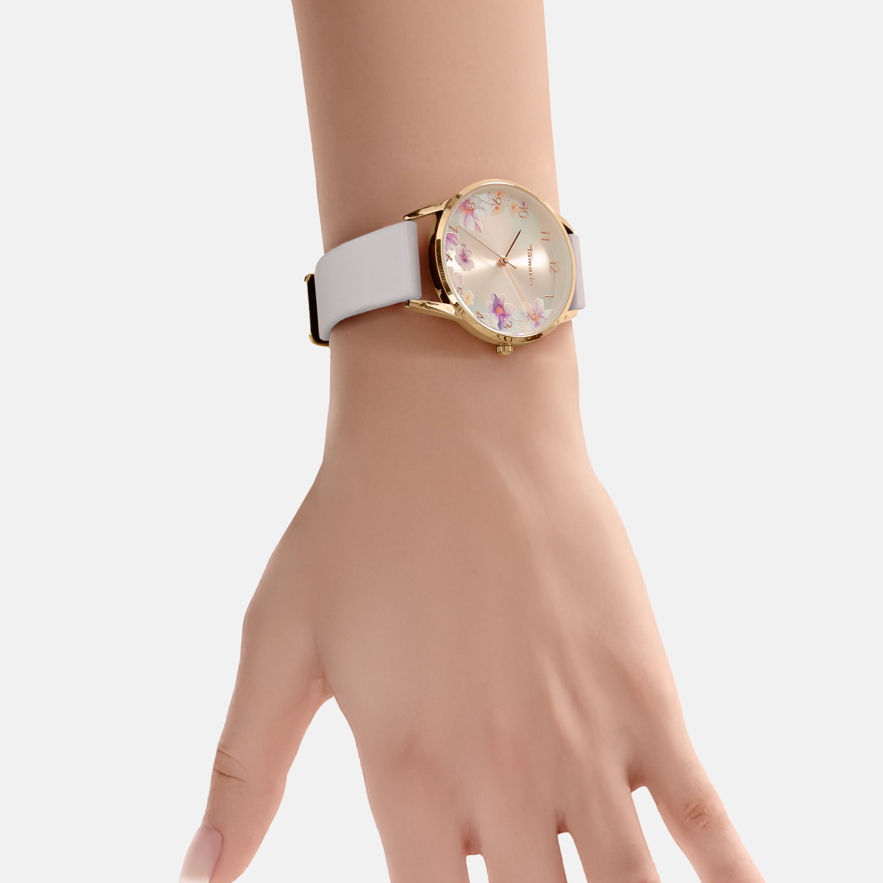 Eine Hand trägt die Tamaris Armbanduhr - The Flower: eine stilvolle Uhr mit Goldgehäuse von Tamaris mit weißem Silikonband und einem eleganten Blumenprint auf dem Zifferblatt.