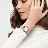 Frau mit langen braunen Haaren in einem weißen Top berührt ihr Haar, während sie die Tamaris Armbanduhr - Sparkling Octavia von Tamaris trägt.