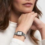 Eine Frau in einem weißen langärmeligen Oberteil, die die Tamaris Armbanduhr - Tiny Watch trägt, stützt ihr Kinn auf ihre Hände - ein Bild von minimalistischer Eleganz.