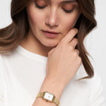 Eine Frau in einem weißen Oberteil, die die Tamaris Armbanduhr - Tiny Watch by Tamaris trägt, schaut nachdenklich mit der Hand am Kinn nach unten - ein Ausdruck minimalistischer Eleganz.