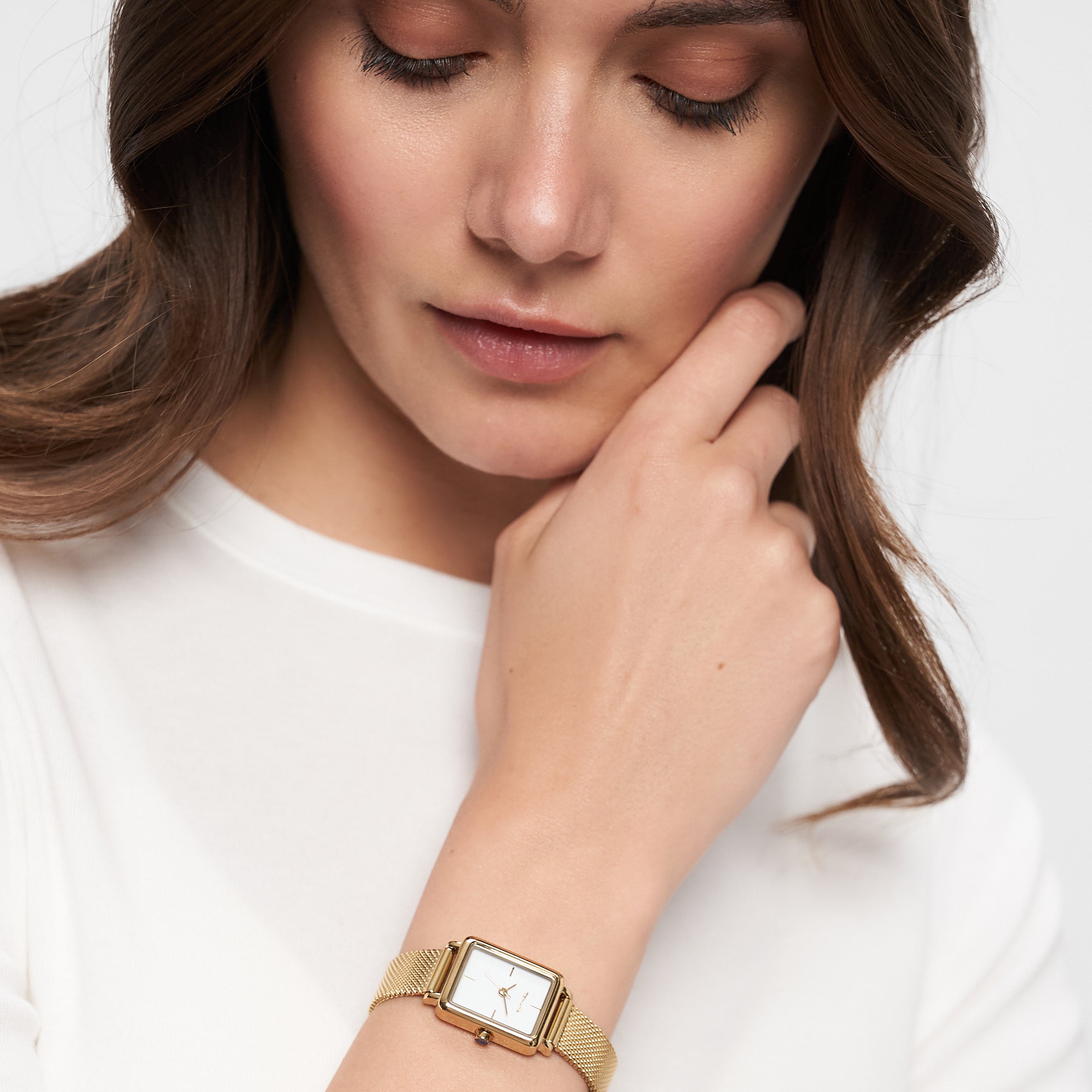 Eine Frau in einem weißen Oberteil, die die Tamaris Armbanduhr - Tiny Watch by Tamaris trägt, schaut nachdenklich mit der Hand am Kinn nach unten - ein Ausdruck minimalistischer Eleganz.