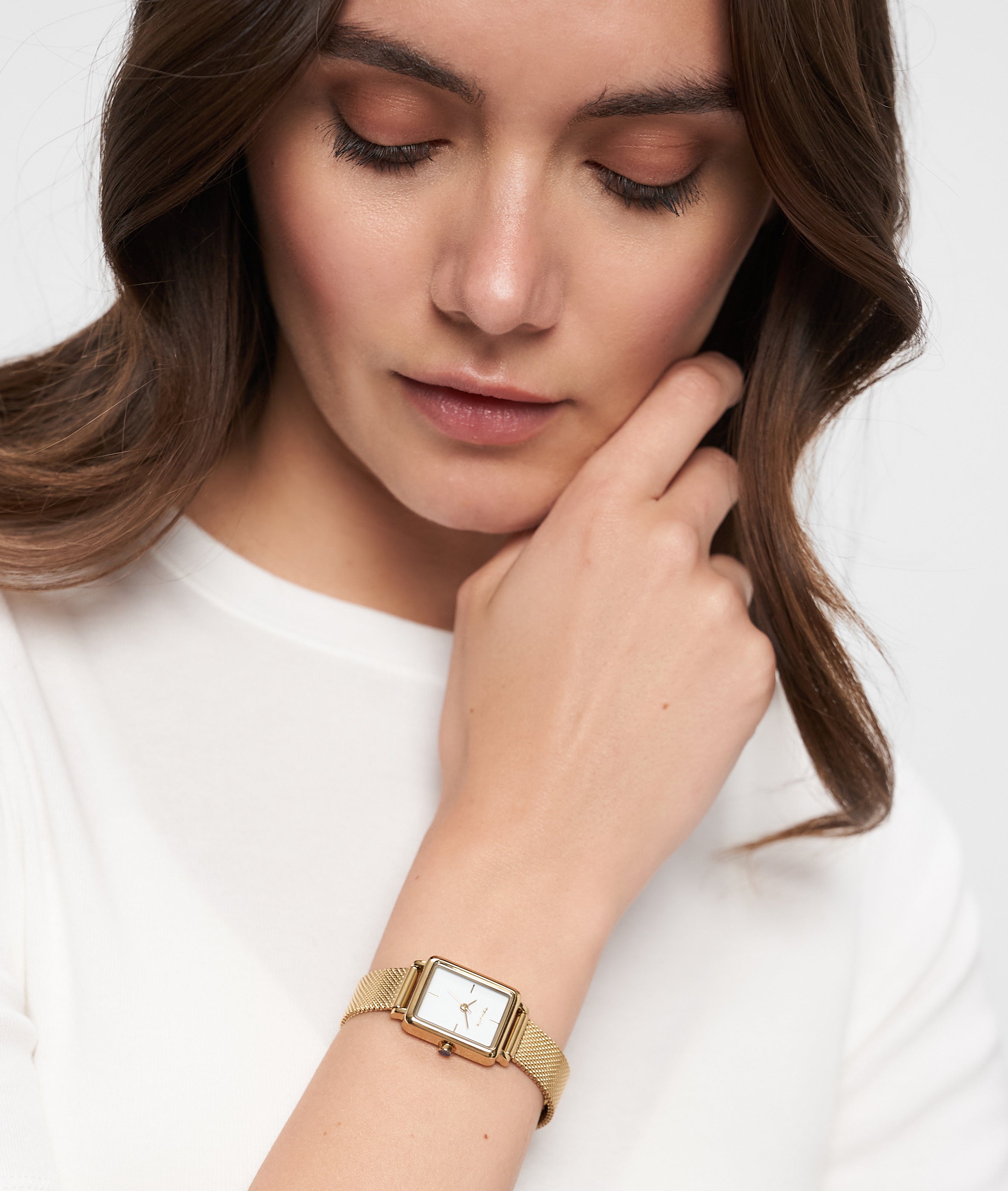Eine Frau in einem weißen Oberteil, die die Tamaris Armbanduhr - Tiny Watch by Tamaris trägt, schaut nachdenklich mit der Hand am Kinn nach unten - ein Ausdruck minimalistischer Eleganz.