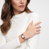 Eine Frau in einem weißen Oberteil blickt auf ihr Handgelenk und trägt die goldene Tamaris Armbanduhr - Tiny Watch von Tamaris, ein Statement von minimalistischer Eleganz.