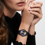 Eine Frau in einem schwarzen Oberteil, die Hände am Kinn verschränkt, trägt die Tamaris Armbanduhr - Sporty Flowers von Tamaris mit ihrem floralen Design und dem sportlichen Silikonband.
