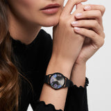 Eine Frau in einem schwarzen Oberteil, die Hände am Kinn verschränkt, trägt die Tamaris Armbanduhr - Sporty Flowers von Tamaris mit ihrem floralen Design und dem sportlichen Silikonband.