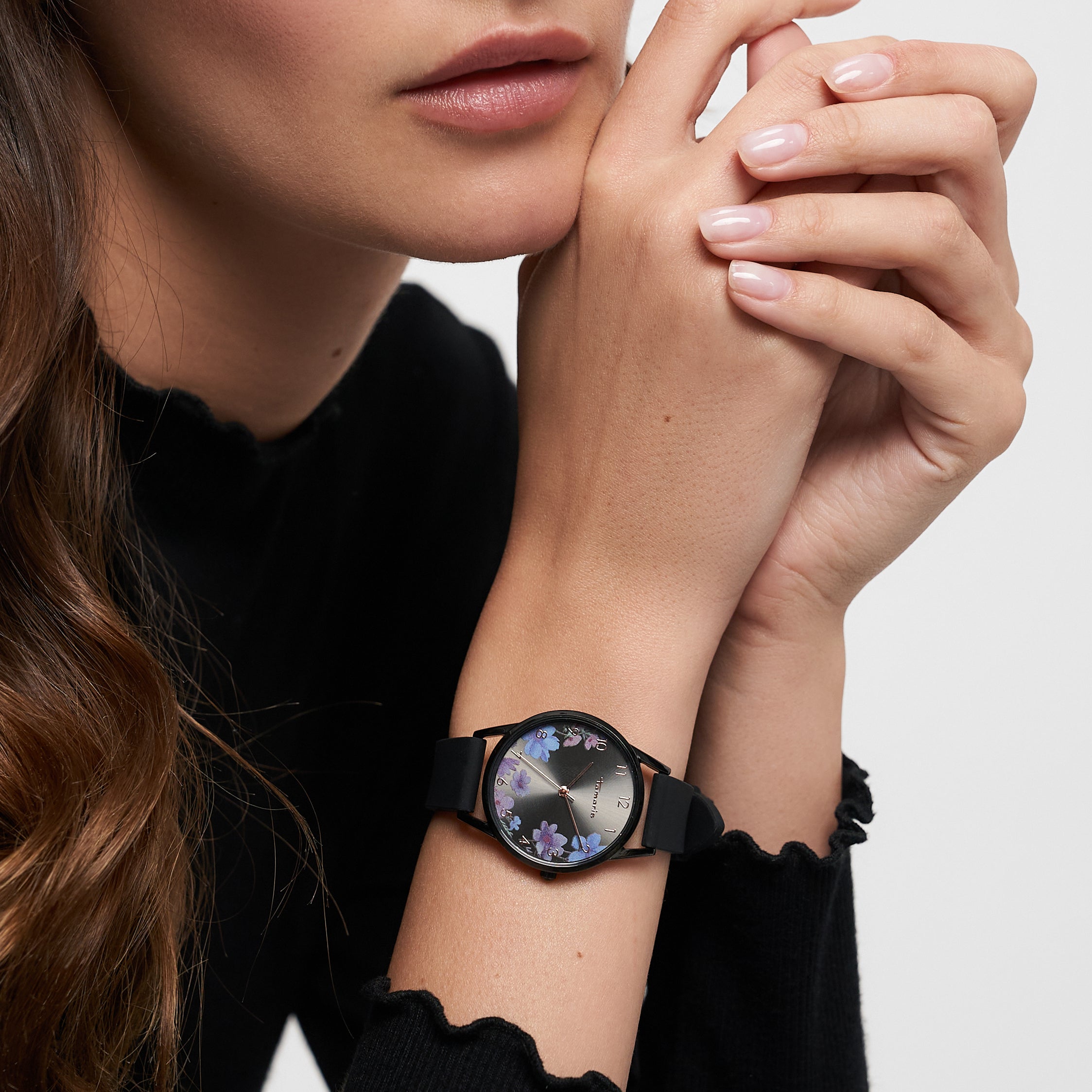 Eine Frau in einem schwarzen Oberteil, die Hände am Kinn verschränkt, trägt die Tamaris Armbanduhr - Sporty Flowers von Tamaris mit ihrem floralen Design und dem sportlichen Silikonband.