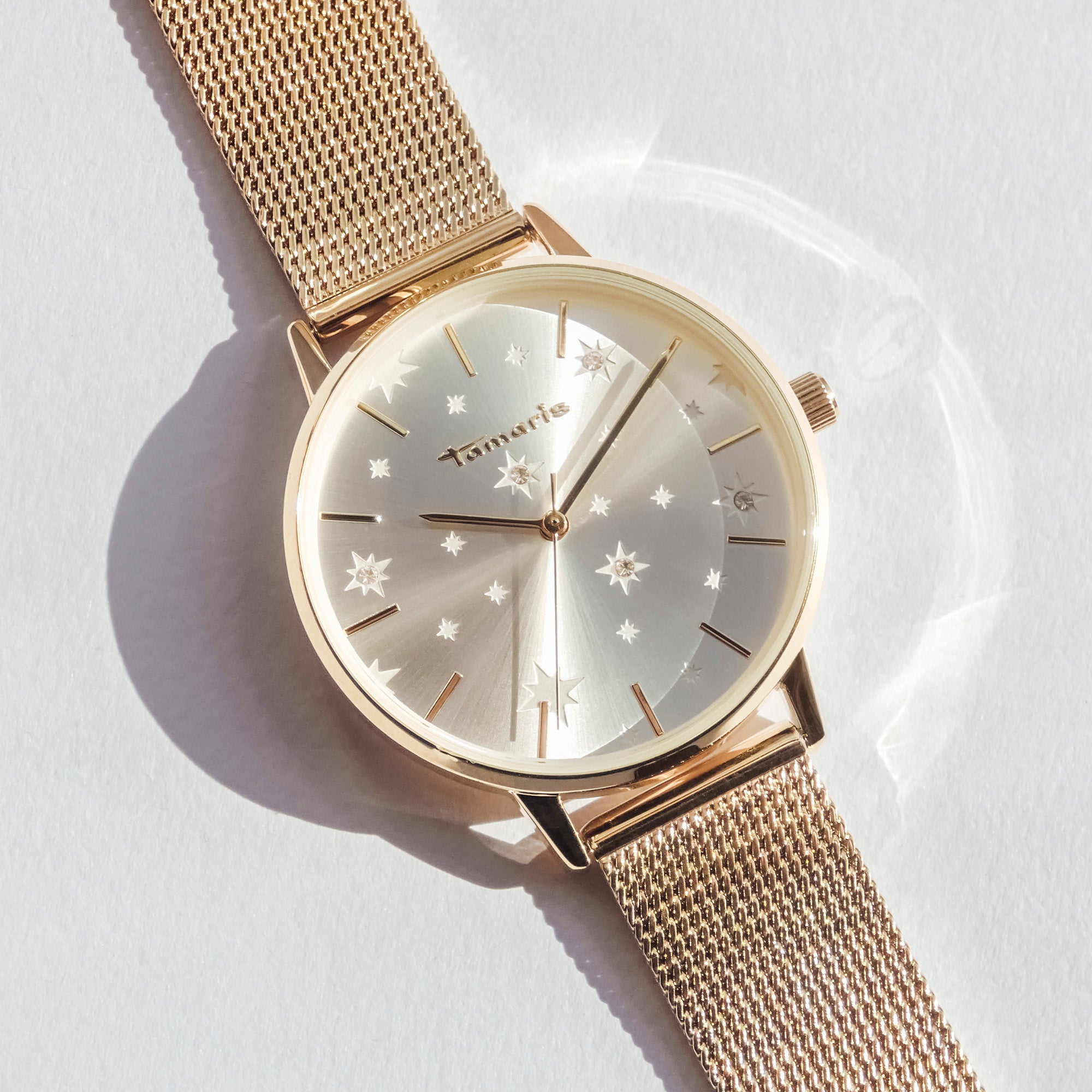 Eine Tamaris Armbanduhr - Shiny Stars mit einem goldenen Mesharmband und einem silbernen Sternenmuster auf der Vorderseite mit funkelnden Zirkoniasteinen, präsentiert auf einer weißen Oberfläche.