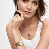 Eine Frau in einem weißen Tank-Top stützt ihr Kinn auf die Hand. Sie trägt die goldene Tamaris Armbanduhr - Shiny Stars, eine goldene Halskette und kleine Creolen-Ohrringe.