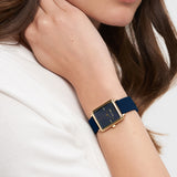 Eine Frau trägt die Tamaris Armbanduhr - The Silicon Square mit einem quadratischen goldenen Gehäuse und einem marineblauen Silikonarmband am linken Handgelenk.