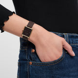 Eine Person trägt ein schwarzes langärmeliges Oberteil, blaue Jeans und eine sportliche Tamaris Armbanduhr - The Silicon Square: eine quadratische schwarze Uhr mit goldenem Rahmen von Tamaris.