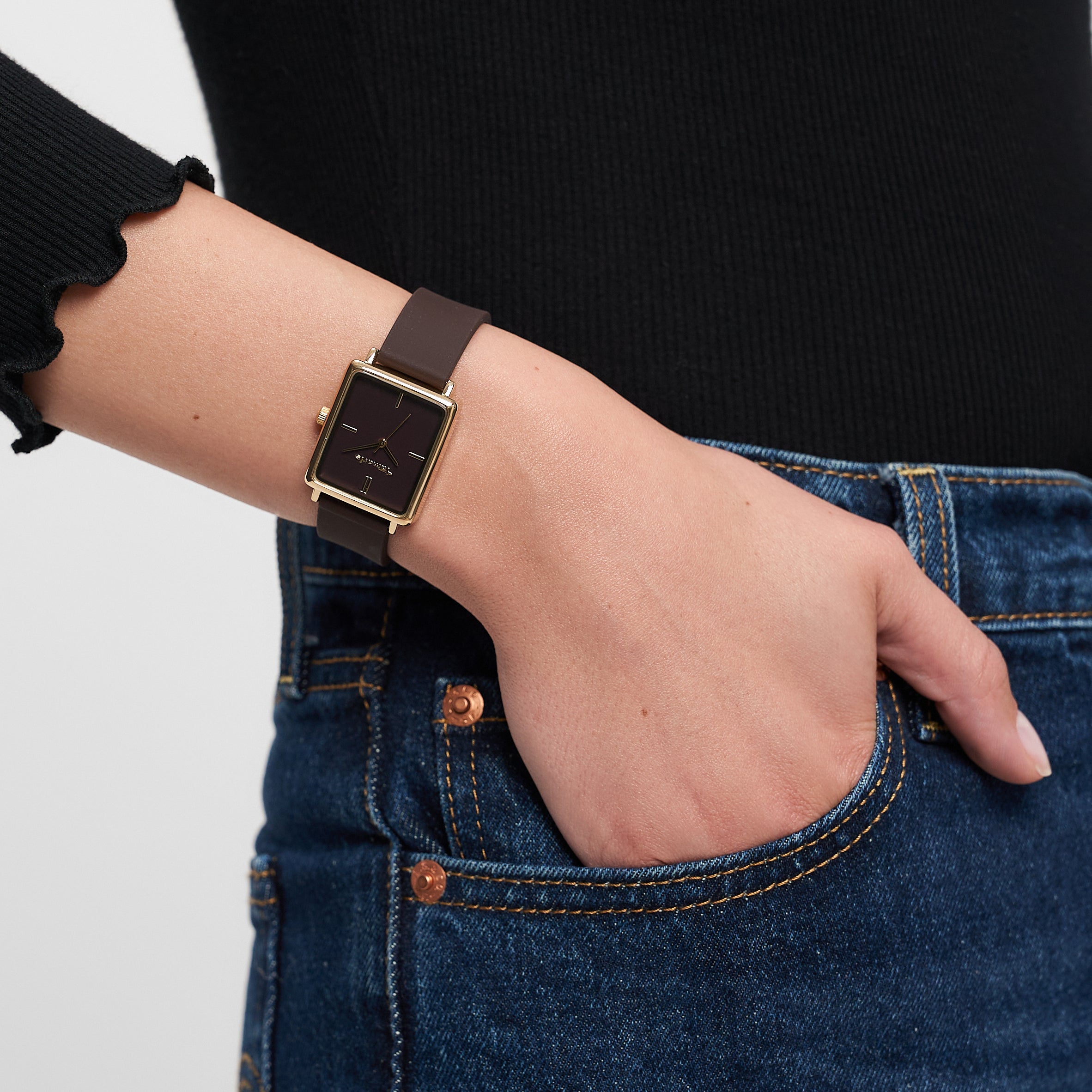 Eine Person trägt ein schwarzes langärmeliges Oberteil, blaue Jeans und eine sportliche Tamaris Armbanduhr - The Silicon Square: eine quadratische schwarze Uhr mit goldenem Rahmen von Tamaris.