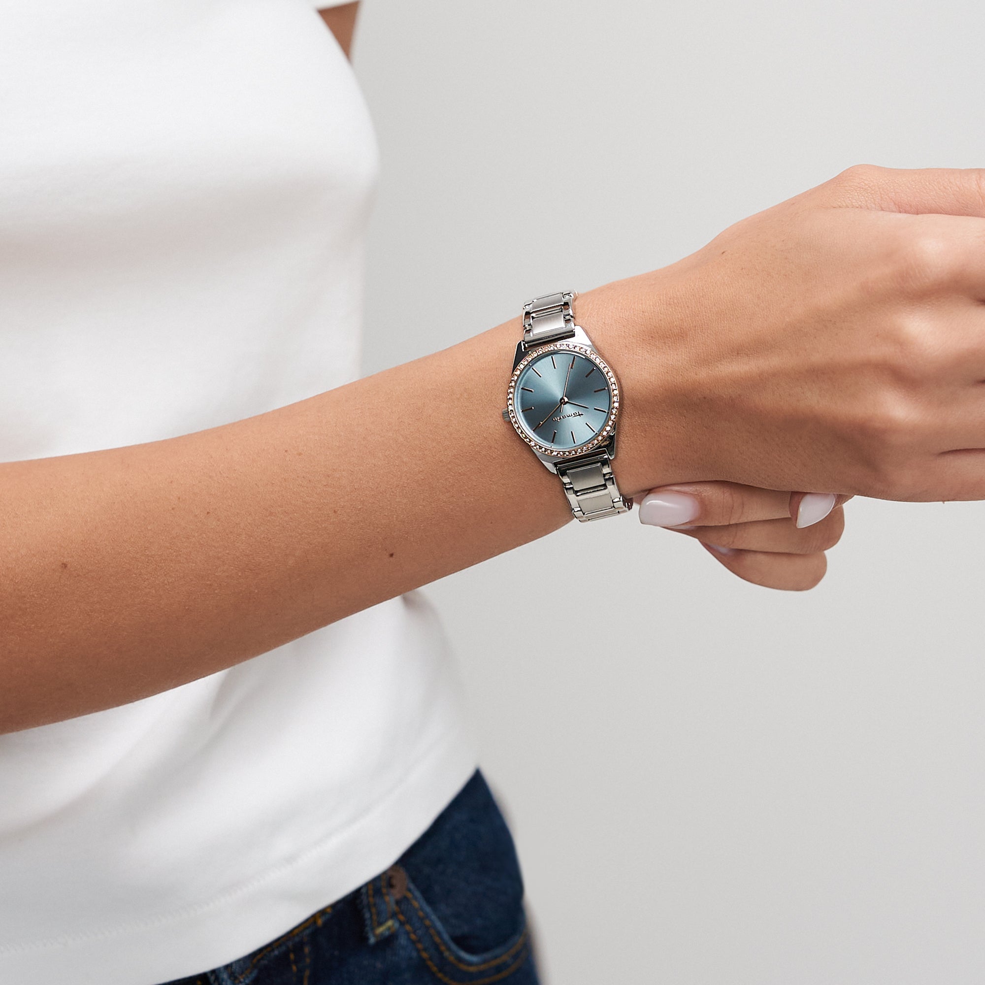 Eine Frau, die ein weißes Oberteil trägt, passt ihre Tamaris Armbanduhr - Modern Classic mit silbernem Zifferblatt und Edelstahlarmband an ihrem linken Handgelenk an.